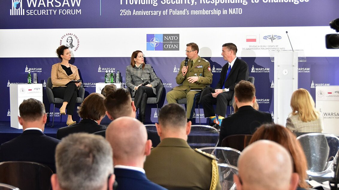 4 osoby siedzą na scenie i rozmawiają. Patrzy na nich publiczność. Baner behind za nimi ma napis "Providing Security, Responding to Challenges. 25th anniversary of Poland's membership in NATO"